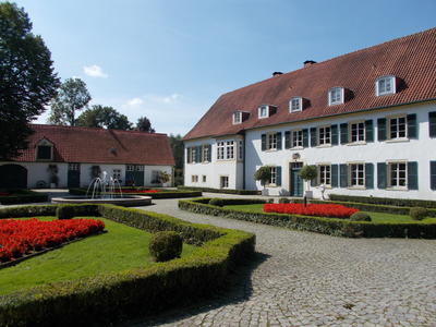 Haus des Gastes: wei�es zweist�ckiges Geb�ude mit gr�nem Fensterl�den, umgeben von Kopfsteinpflaster, formalen Hecken und roten Blumenbeeten unter blauem Himmel