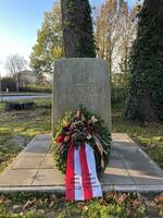 Gedenkstein an einem Wegesrand; davor liegt ein Blumenkranz mit rot-wei�em Trauerband.