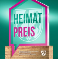 Eine gl�serne Troph�e in Hausform mit pinkem Rahmen und der Aufschrift �HEIMATPREIS� steht auf einem h�lzernen Sockel mit dem Wappen und Schriftzug des nordrhein-westf�lischen Heimatministeriums, vor gr�nem Hintergrund.