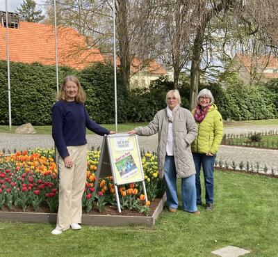 Das Bild zeigt Touristikleiterin Judith Fidler, Regina Titkemeyer vom SuS Holzhausen und Marianne Nolte vom nwwe e.V. im Kurpark vor dem Haus des Gastes. In ihrer Mitte steht ein Aufsteller, der auf das Aktivangebot "Sport im Park" hinweist.