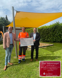 B�rgermeister Marko Steiner, Bauhofleiter Daniel Pluntke und Gesch�ftsstellenleiter Bernd Rust stehen unter einem Sonnensegel auf einem Spielplatz und halten einen Scheck in der Hand.
