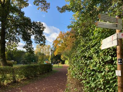 Im Vordergrund steht der Weg, der weiter in den Kurpark hinein geht. Dieser besteht aus rotem Schotter, der Weg wird begleitet von gr�nen Hecken und B�umen mit einem orangenen Blattwerk. Auf der rechten Seite des Bildes steht noch eine Wandermarkierung mit Pfeilen in den die verschiedenen Routen gehen