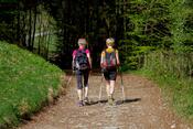 Zu sehen sind zwei Frauen, die eine mit einem gelben T-Shirt und die andere, mit einem pinken T-Shirt. Beide haben Gehst�cke in der Hand und wandern in eine kleine Waldpassage rein