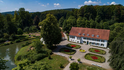 Drohnenaufnahme von dem Haus des Gastes und dem anliegendem Kurpark an einem Sommertag mit klarem blauen Himmel. Das Haus, sowie der Kurpark sind von vielen gro�en gr�nen B�umen umgeben