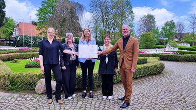 Zu sehen sind von links nach rechts: Judith Fidler (Touristikleitung), Britta B�ker und Christina Hoppe (beide Bezirksregierung Detmold), Gabriele Schnabel (Touristikverein Bad Holzhausen) und B�rgermeister Marko Steiner bei der Urkunden�bergabe vor der bl�henden Kulisse des Kurparks Bad Holzhausen.